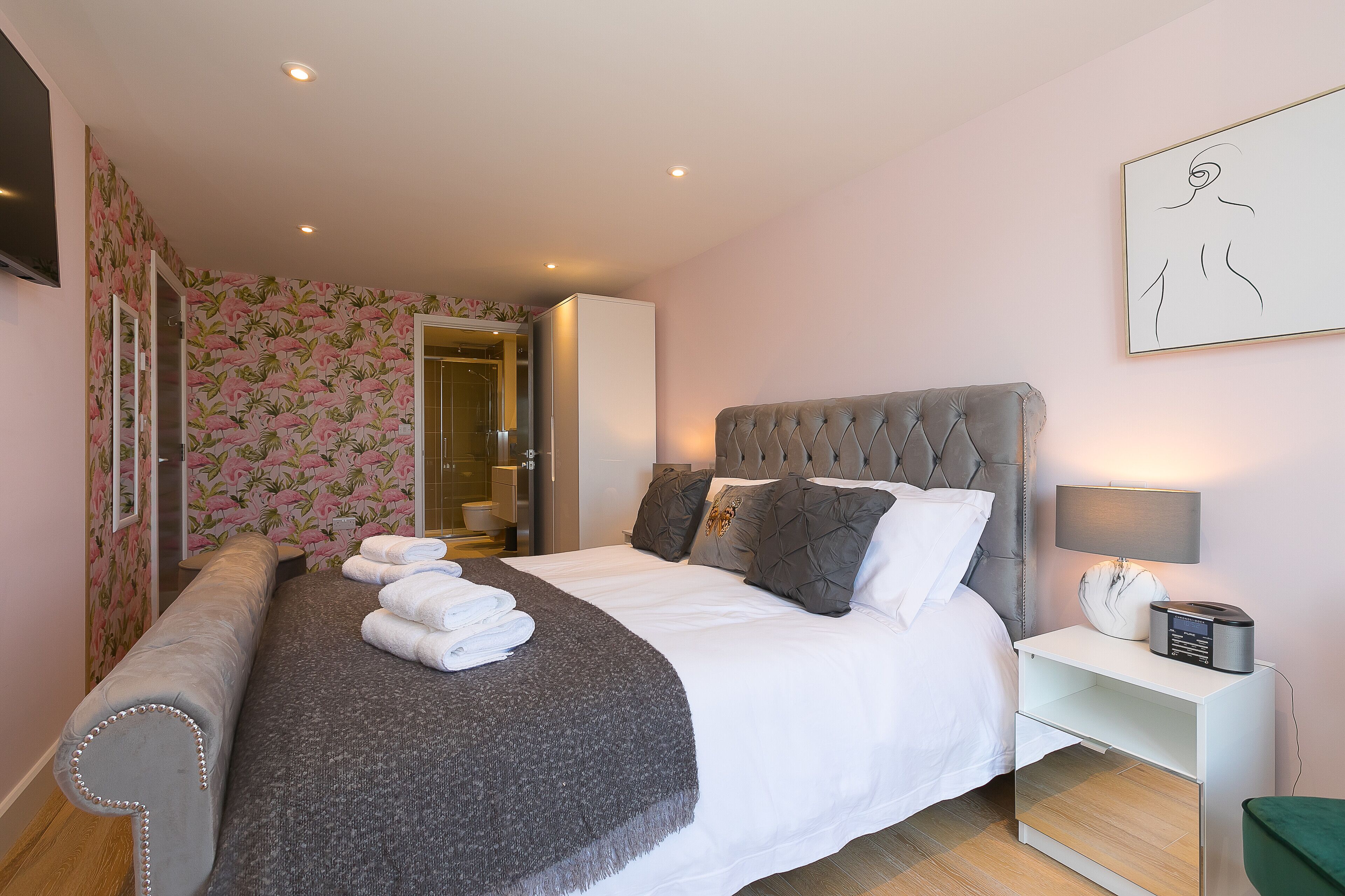 Een netjes opgemaakt bed met een grijze hoofdbord, een nachtkastje met een lamp, en een ingelijste afbeelding aan de muur.