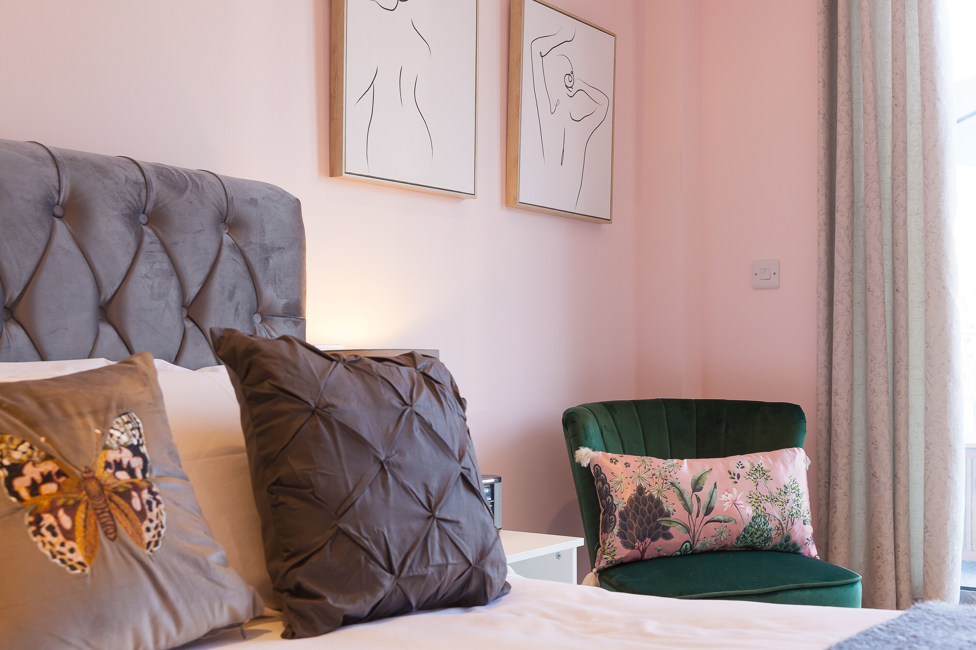 Ein Schlafzimmer mit einem gesteppten Kopfteil, einem Bett mit vielen Kissen, einem grünen Sessel und zwei gerahmten Kunstwerken an der Wand.