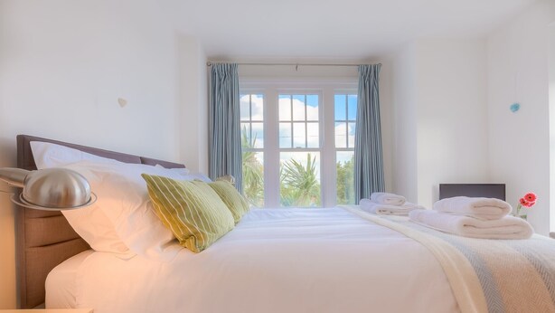 A neatly made bed with white linens and a green pillow, a bedside lamp, and a window with curtains. - Sea Dream (St Ives)