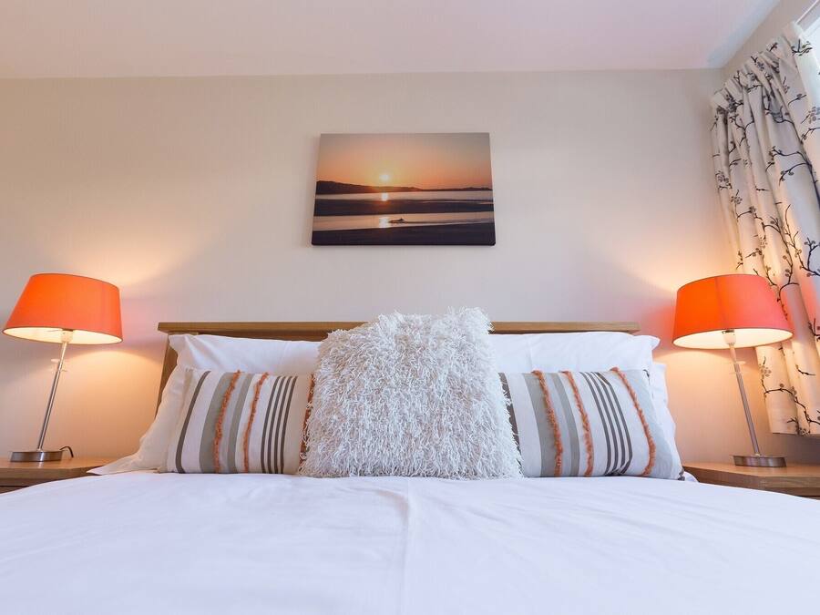 Una cama bien hecha con sábanas blancas y almohadas a rayas, flanqueada por dos lámparas de noche con pantallas naranjas, y un cuadro con un atardecer sobre la cama.