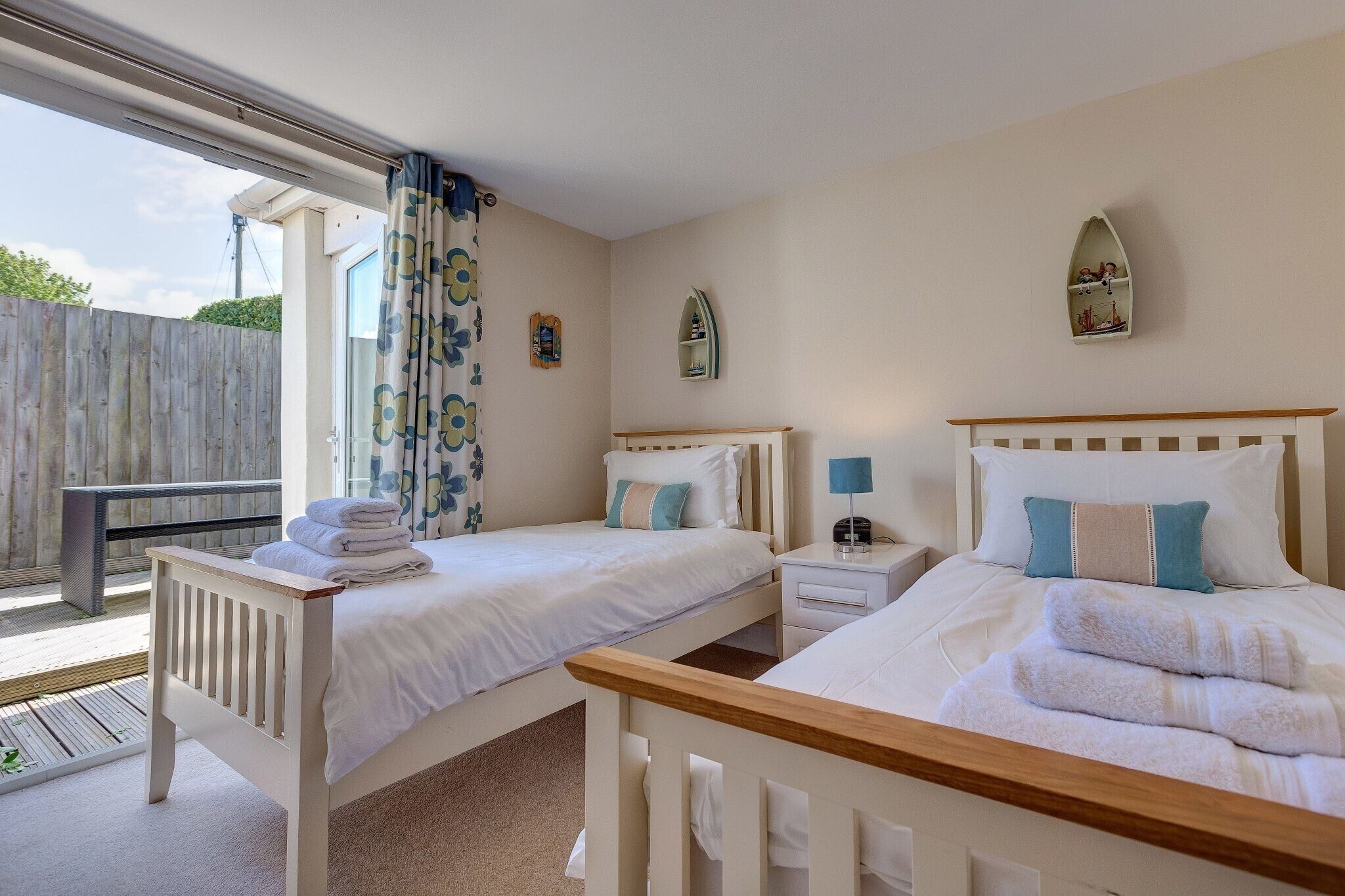 Une chambre avec deux lits, une petite table de chevet avec une lampe, une fenêtre avec des rideaux et une terrasse en bois à l’extérieur.