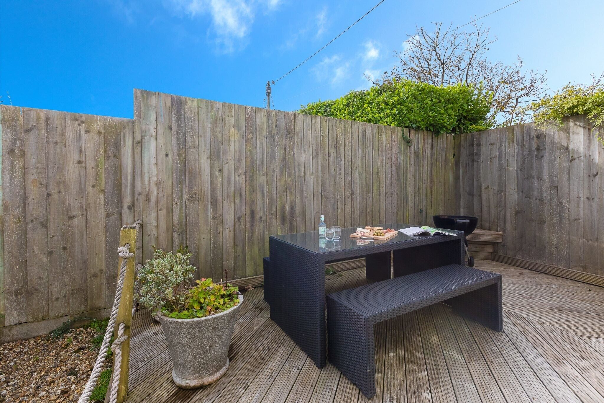 A wooden deck with a glass-top table and a bench, surrounded by a wooden fence and potted plants.