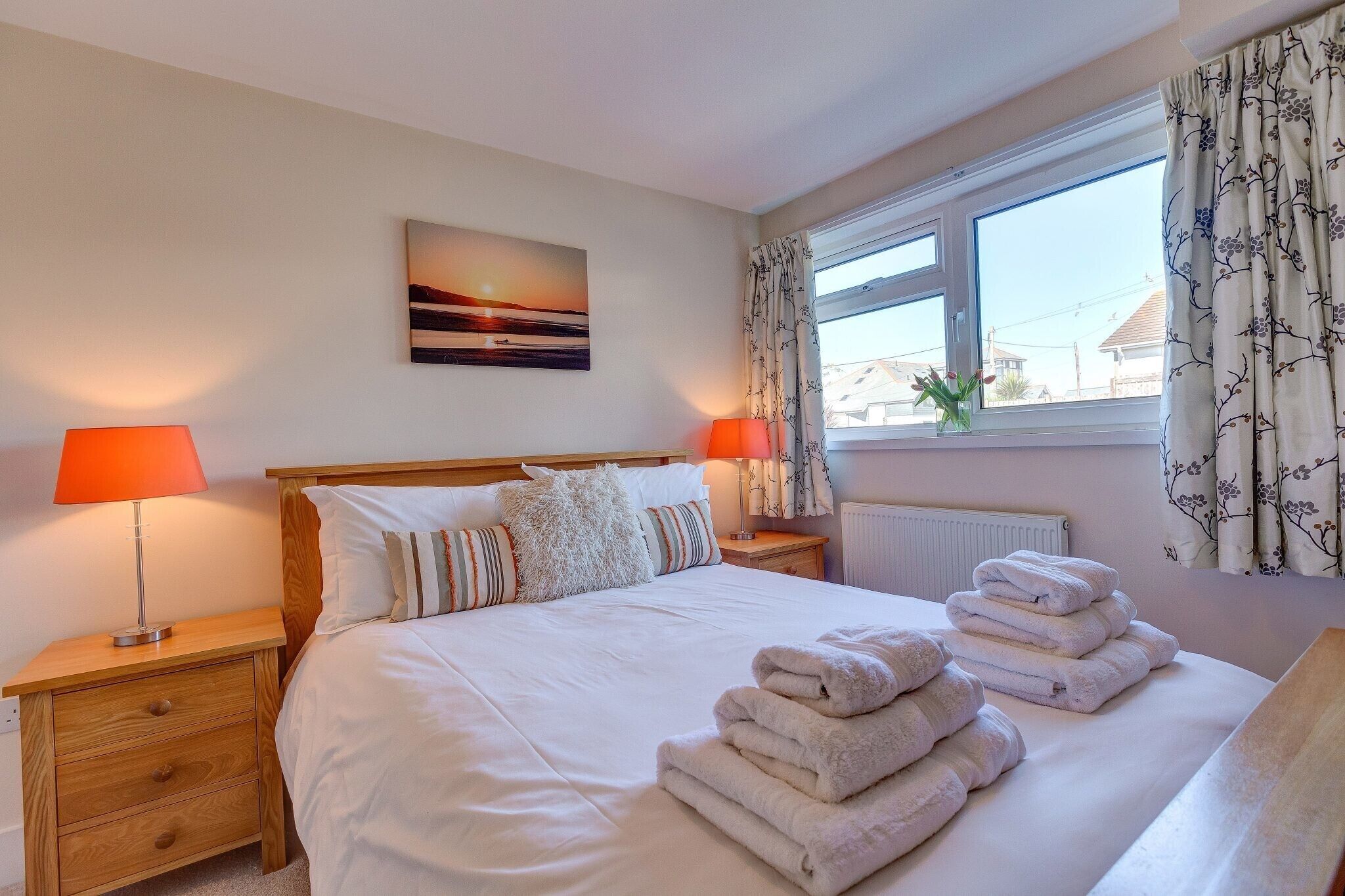 A neatly made bed with white linens and a stack of towels, two bedside lamps, a window with floral curtains, and a sunset painting on the wall.