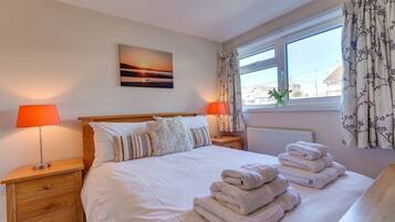 A neatly made bed with white linens and a stack of towels, two bedside lamps, a window with floral curtains, and a sunset painting on the wall.