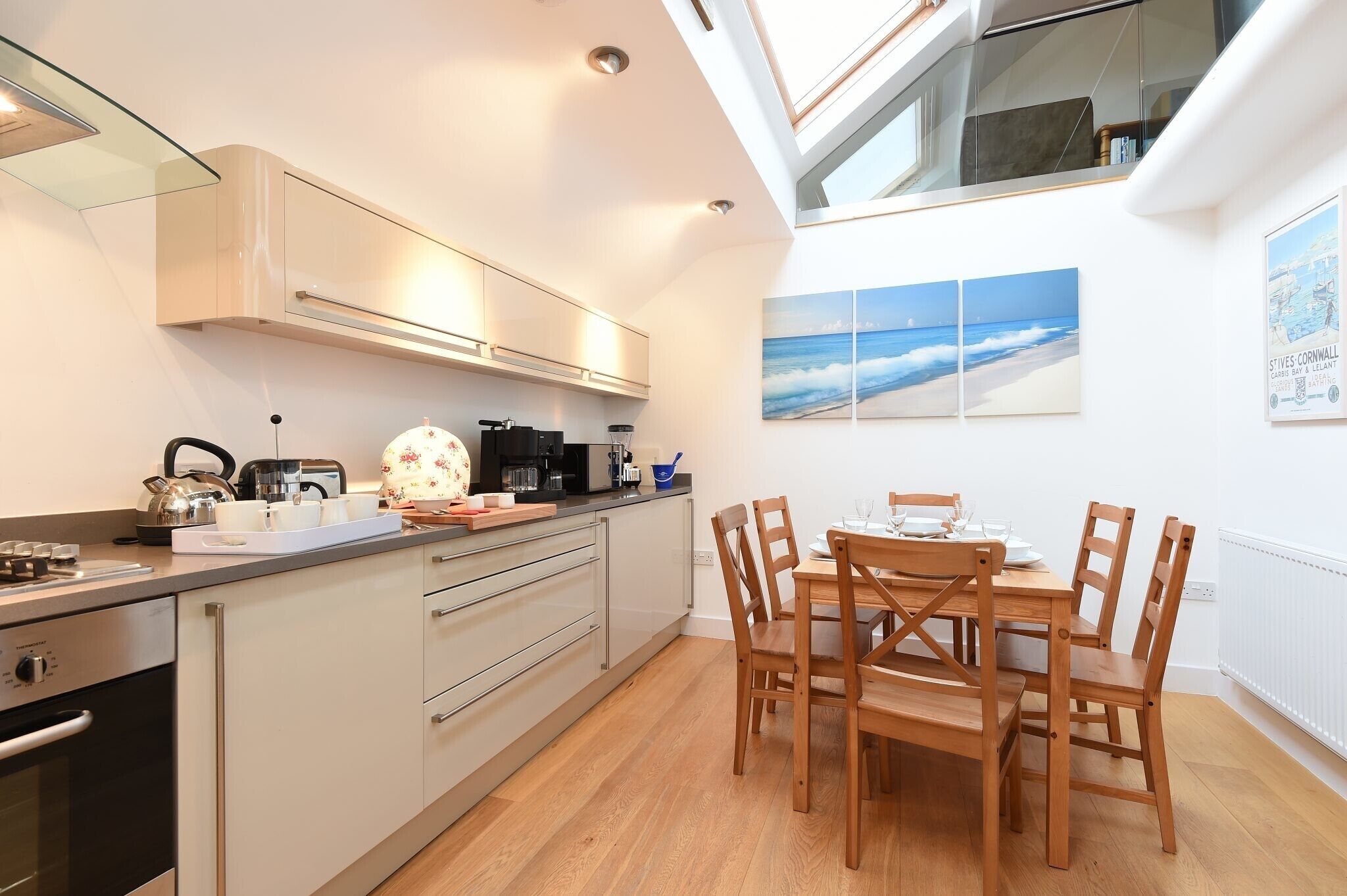 A modern kitchen with a dining area, featuring a wooden table and chairs, a built-in oven, and a view of the sea through the window.