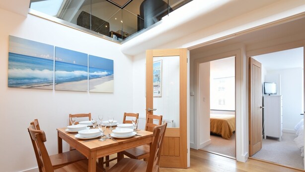 A dining area with a wooden table set for four, a large beach mural on the wall, and a view of the bedroom through an open door.