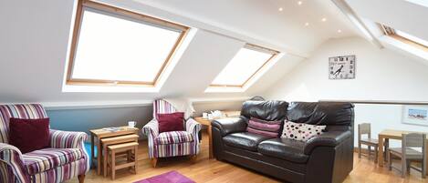 A loft-style living room with a high ceiling, skylights, a black leather sofa, striped armchairs, a small wooden table, and a purple rug.