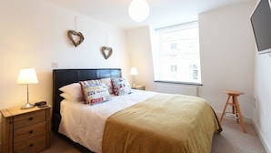 A bedroom with a bed, two heart-shaped wall decorations, bedside lamps, a wooden nightstand, a window with a view, and a TV mounted on the wall.