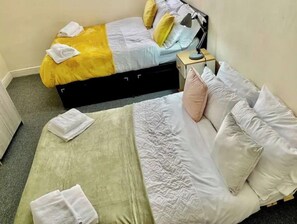 A bedroom with two beds, one with a yellow and blue bedspread and the other with a green and white bedspread. There is a nightstand with a lamp and a box on it.