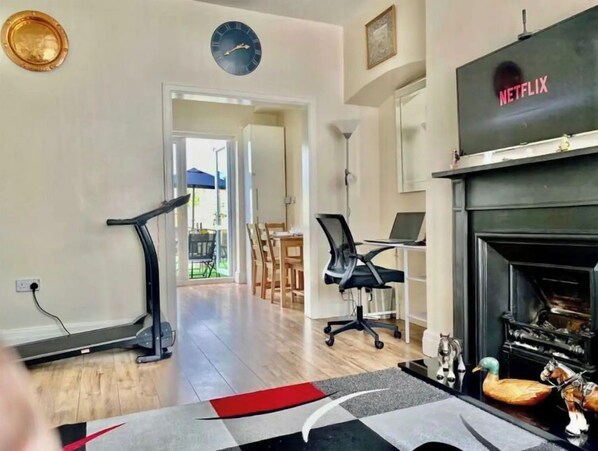 A living room with a treadmill, a desk with a laptop, a fireplace, and a clock on the wall.