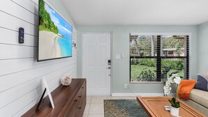 A modern living room with a flat-screen TV mounted on the wall, a wooden coffee table, a sofa, and a window with a view of greenery. - "Flamingo Feliz" Heated Pool! Close to Beach and 5th Ave. (Naples)