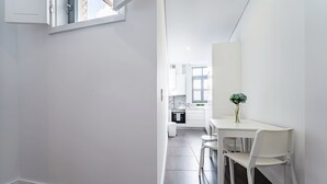 Interior modern i blanc amb zona de dinar que inclou taula i cadires, un vas amb una planta, i cuina al fons.