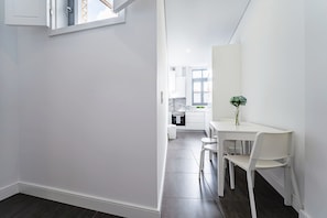 A modern, white interior with a dining area featuring a table and chairs, a vase with a plant, and a kitchen in the background. - Nomad's Formosa Harmony - 1BED Porto (Porto)