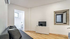 Un saló modern amb un sofà gris, un televisor de pantalla plana sobre un moble blanc, terres de fusta i una finestra amb vistes.
