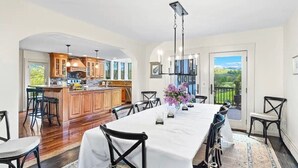 A spacious dining area with a long table, black chairs, and a white tablecloth. There is a kitchen area with wooden cabinets and a view of the outdoors through large windows.