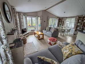 Ein geräumiges Wohnzimmer mit Sofa, Couchtisch und Fernseher. Große Fenster mit Vorhängen, ein Essbereich und ein Bücherregal sind ebenfalls vorhanden.