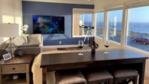 Un salón moderno con un ventanal grande con vista al océano, una mesa de comedor con copas de vino, un telescopio y un cuadro grande en la pared.