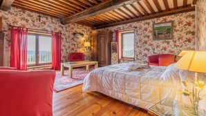 Un dormitorio con techo de madera, papel tapiz floral, una cama, un sillón rojo, una mesa de madera y una ventana con vistas.