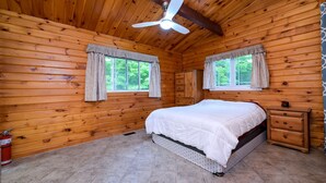 A wooden cabin bedroom with a bed, a dresser, a ceiling fan, and two windows with curtains.