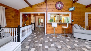 A log cabin interior with a checkered floor, a wooden ceiling, a clock, a kitchen area with a sink and cabinets, and a staircase.