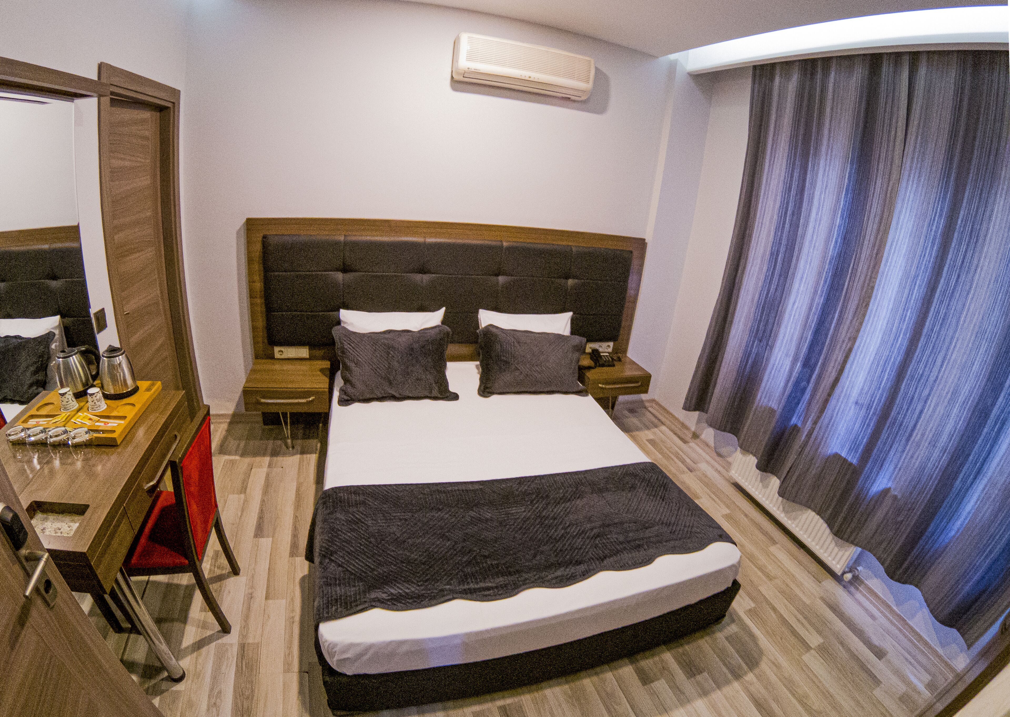 A hotel room with a large bed, bedside tables, a desk with a chair, a mirror, and a window with curtains.