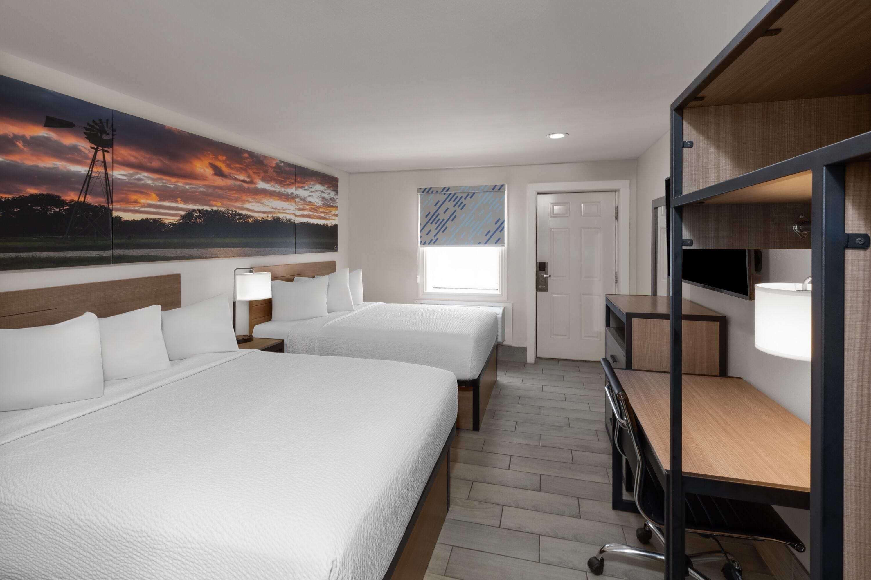 A hotel room with two beds, a desk, a TV, and a painting of a sunset.
