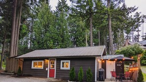 A small cabin with a red door, a porch with outdoor seating, and a gravel driveway.