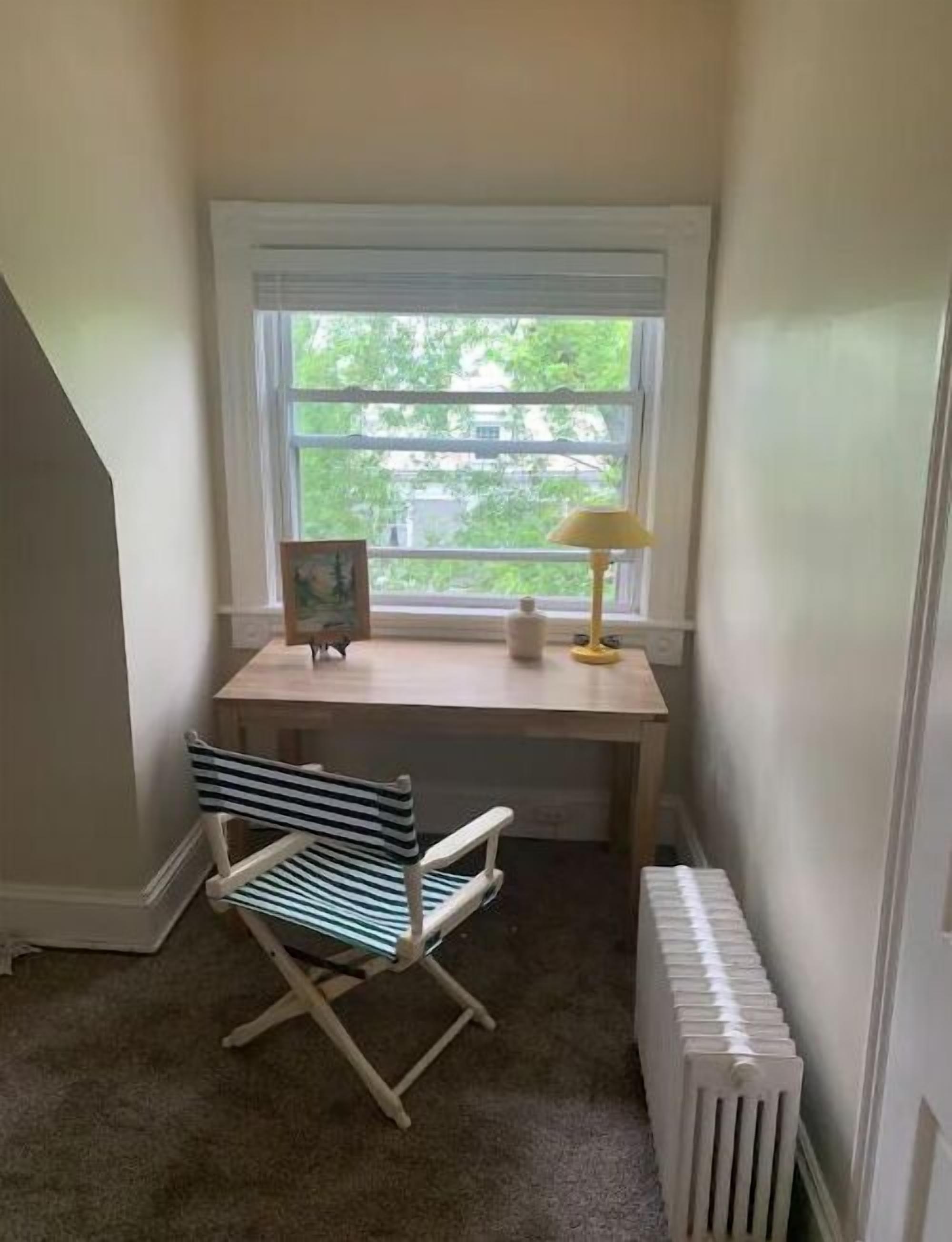 A corner room with a window, a desk, a chair, a radiator, and a picture frame.