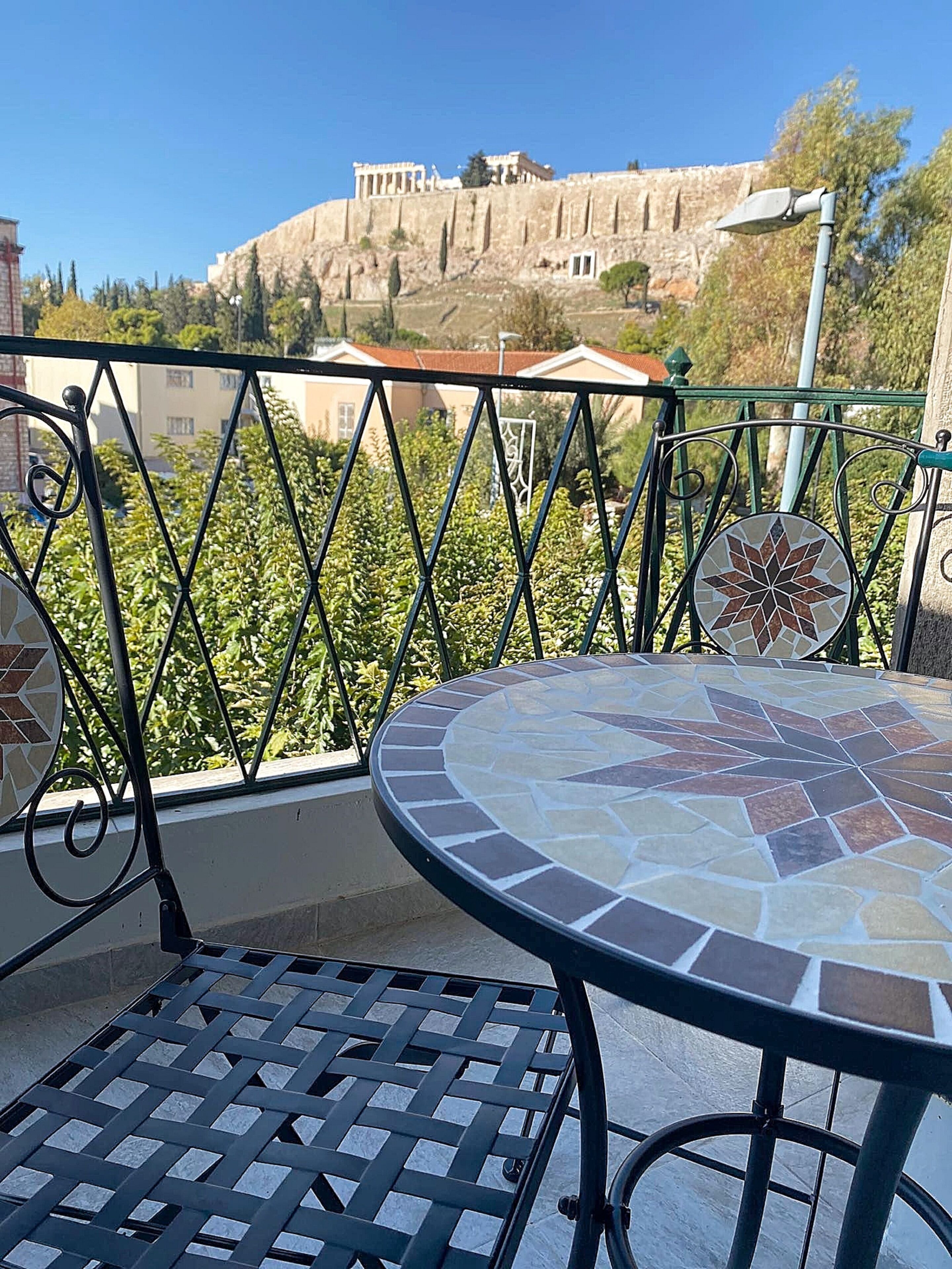 A balcony with a round table and metal grill, overlooking a historic building with columns and a stone wall.