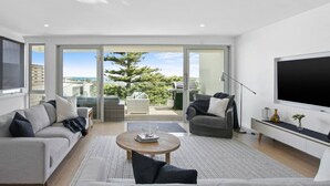 A modern living room with a sofa, armchair, and coffee table, a flat-screen TV, and a sliding glass door leading to a balcony with outdoor furniture. - Beachside Living with Sea Views at the Pines (Ocean Grove)