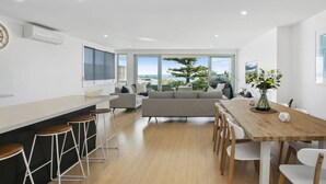 A modern kitchen with a large island, bar stools, and a dining area with a wooden table and chairs. - Beachside Living with Sea Views at the Pines (Ocean Grove)