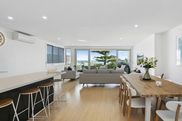 A modern kitchen with a large island, bar stools, and a dining area with a wooden table and chairs. - Beachside Living with Sea Views at the Pines (Ocean Grove)