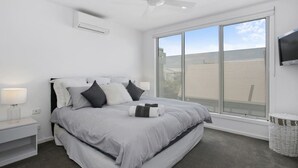 A modern bedroom with a large bed, a ceiling fan, a window with blinds, and a TV mounted on the wall. - Beachside Living with Sea Views at the Pines (Ocean Grove)