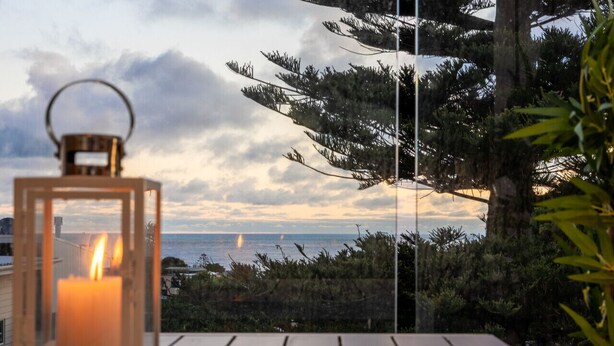 A candle in a lantern on a wooden table with a view of the sea and trees. - Beachside Living with Sea Views at the Pines (Ocean Grove)