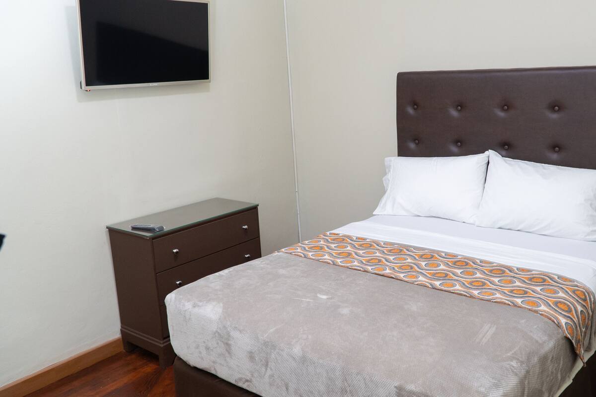 A neatly arranged hotel room with a bed, a bedside table, a TV mounted on the wall, and a small nightstand with a lamp.