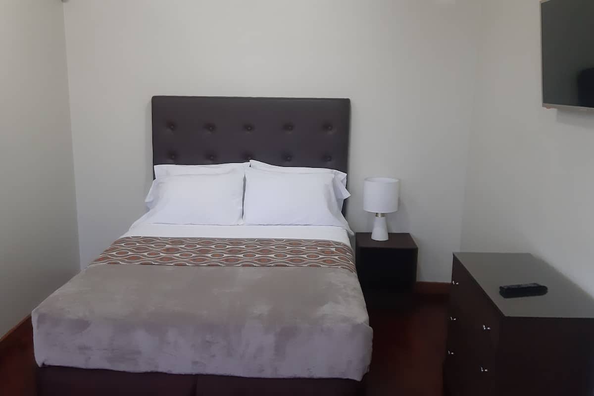 A neatly made bed with a patterned bedspread and white pillows, a bedside table with a lamp, and a small cabinet.