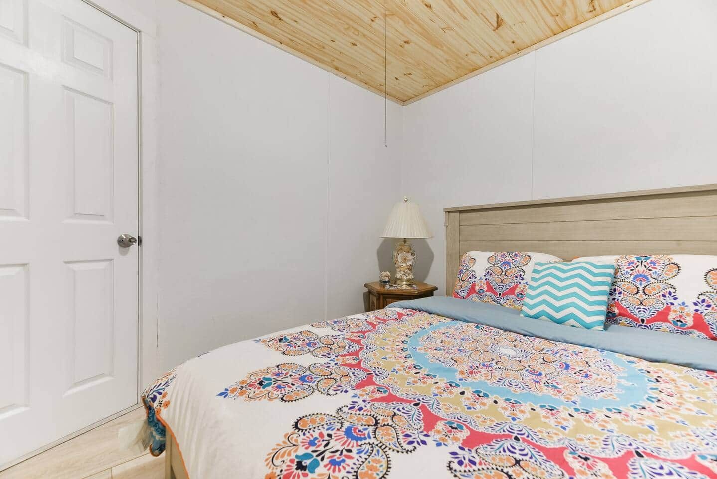 Ein Schlafzimmer mit Holzboden, einer weißen Tür, einem Bett mit gemusterten Betttextilien, einem Nachttisch mit Lampe und einem Zierkissen.