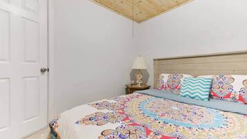 Un dormitorio con techo de madera, una puerta blanca, una cama con ropa de cama estampada, una mesita de noche con una lámpara y un cojín decorativo.