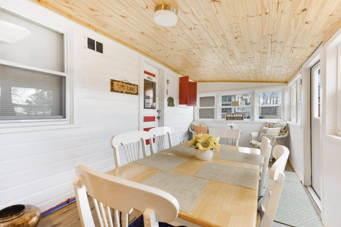 A cozy cabin interior with a wooden ceiling, a dining table with chairs, and a window with a 'WELCOME TO THE COTTAGE' sign.