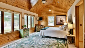 A log cabin bedroom with a large bed, a dresser, a TV, and a painting of a deer. - Private Lake Resort: large yard, sandy beach, pontoon, fire pit, home theater (Spooner)