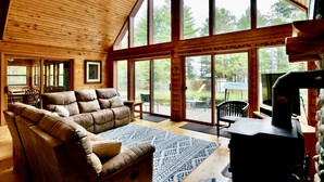 A spacious living room with a large wooden ceiling, a leather sofa, a dining table with chairs, and a grill outside the large windows. - Private Lake Resort: large yard, sandy beach, pontoon, fire pit, home theater (Spooner)