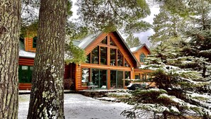 A cabin with a large glass front, surrounded by snow-covered trees. - Private Lake Resort: large yard, sandy beach, pontoon, fire pit, home theater (Spooner)