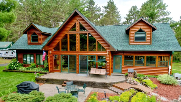 A log cabin with a large front porch, a covered patio, and a landscaped garden. - Private Lake Resort: large yard, sandy beach, pontoon, fire pit, home theater (Spooner)