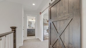 Un pasillo con puerta de madera, suelo alfombrado y una escalera con barandilla de metal.