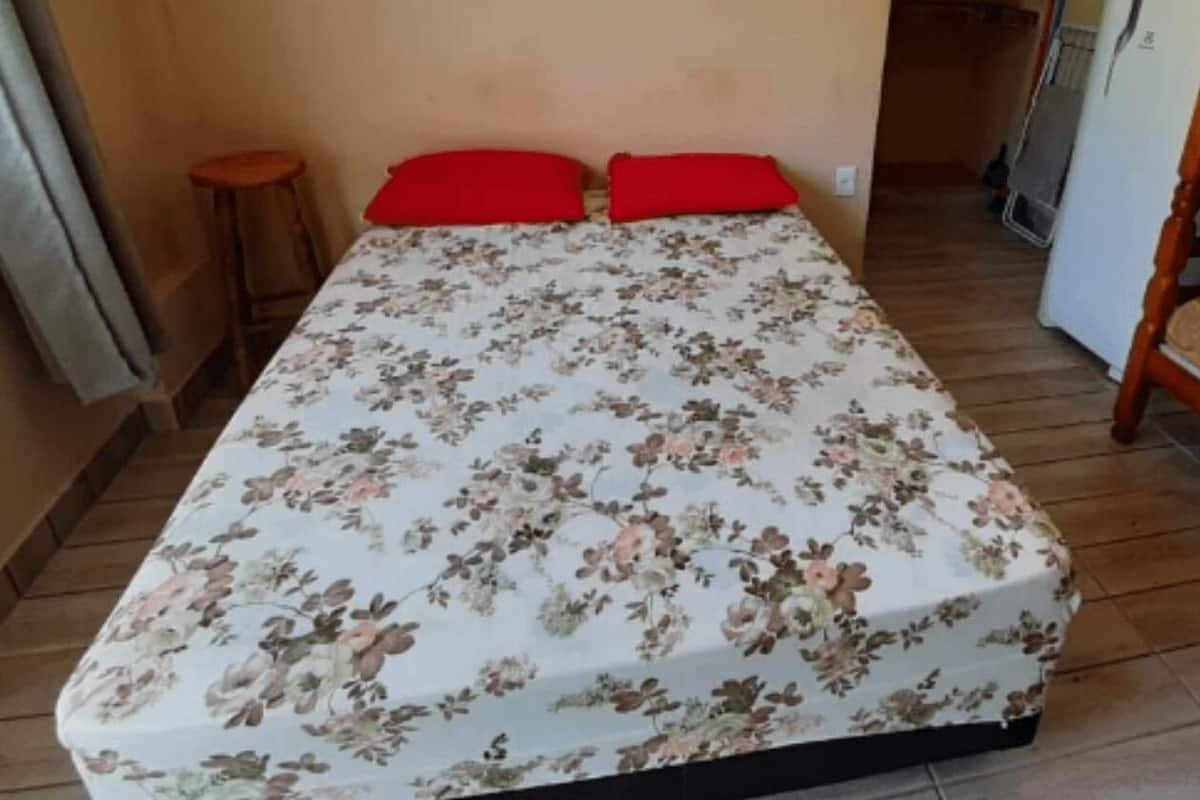 A single bed with a floral bedspread and two red pillows, a wooden bench, and a small stool in a room with beige walls and a tiled floor.
