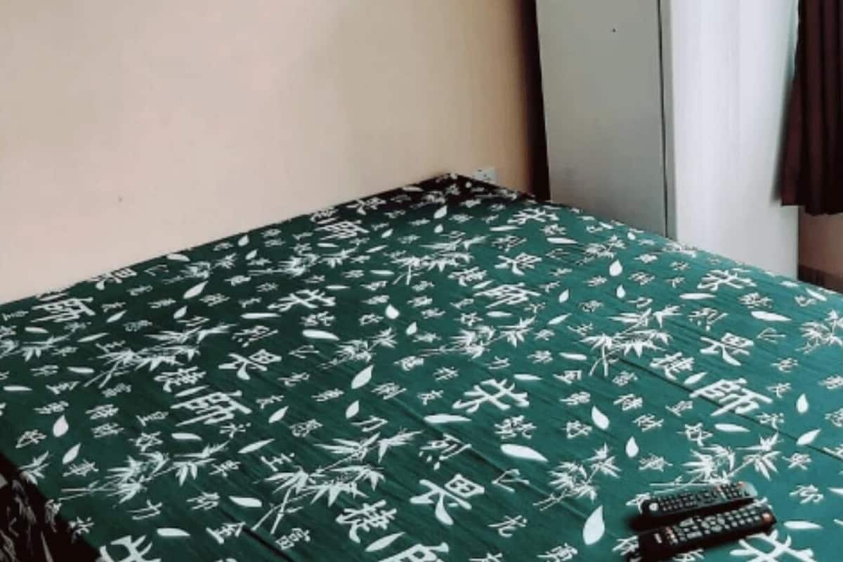 A room with a bed covered in a green bedspread with white patterns, a standing fan, and a window with curtains.