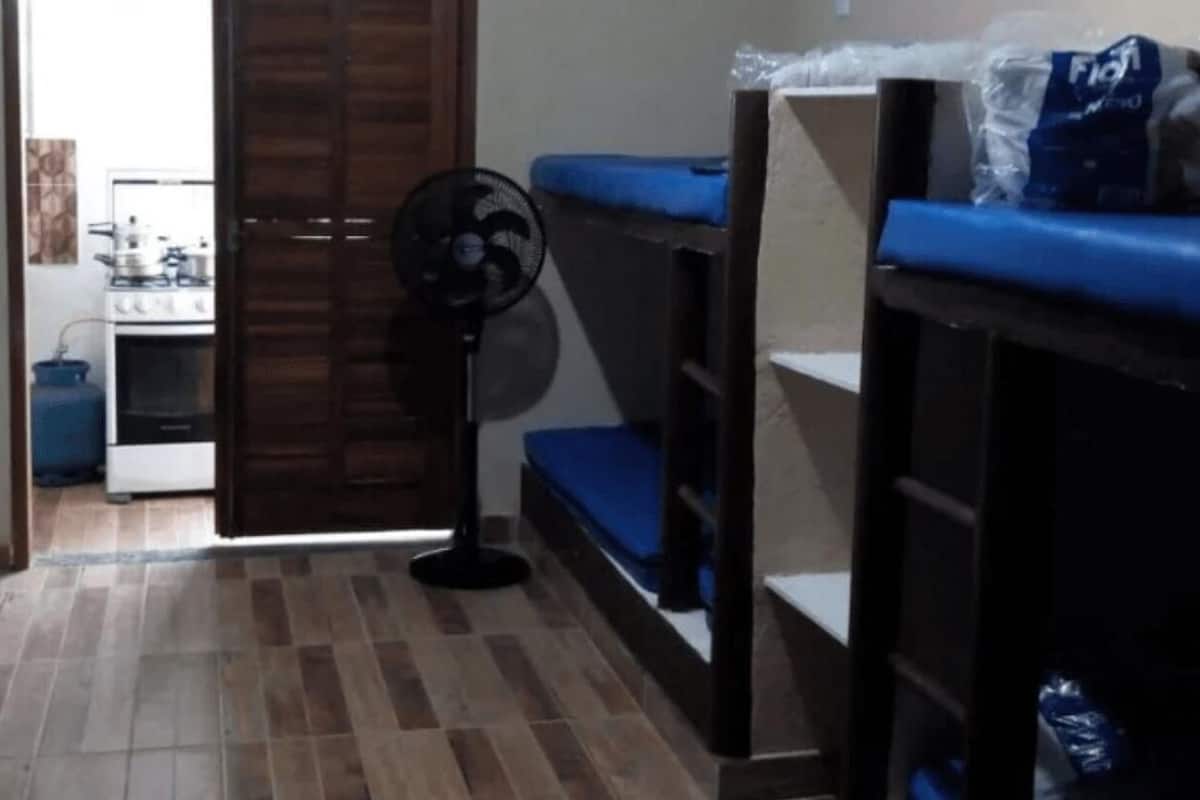 A dormitory room with bunk beds, a fan, and a kitchenette in the background.
