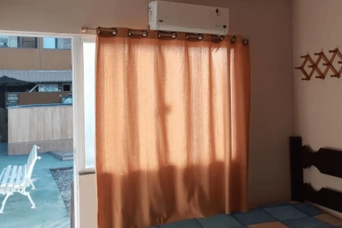 A room with a bed, a window with curtains, a wall-mounted heater, and a small table with a printer.