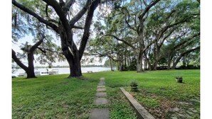 A pathway leading through a park with large trees and a body of water in the background.
