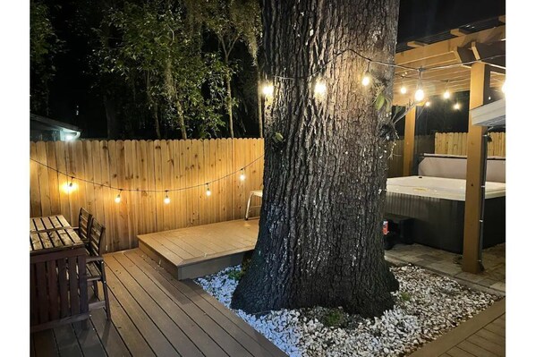 Relax in a serene outdoor space with a wooden deck, tree, and string lights, perfect for dining or unwinding.
